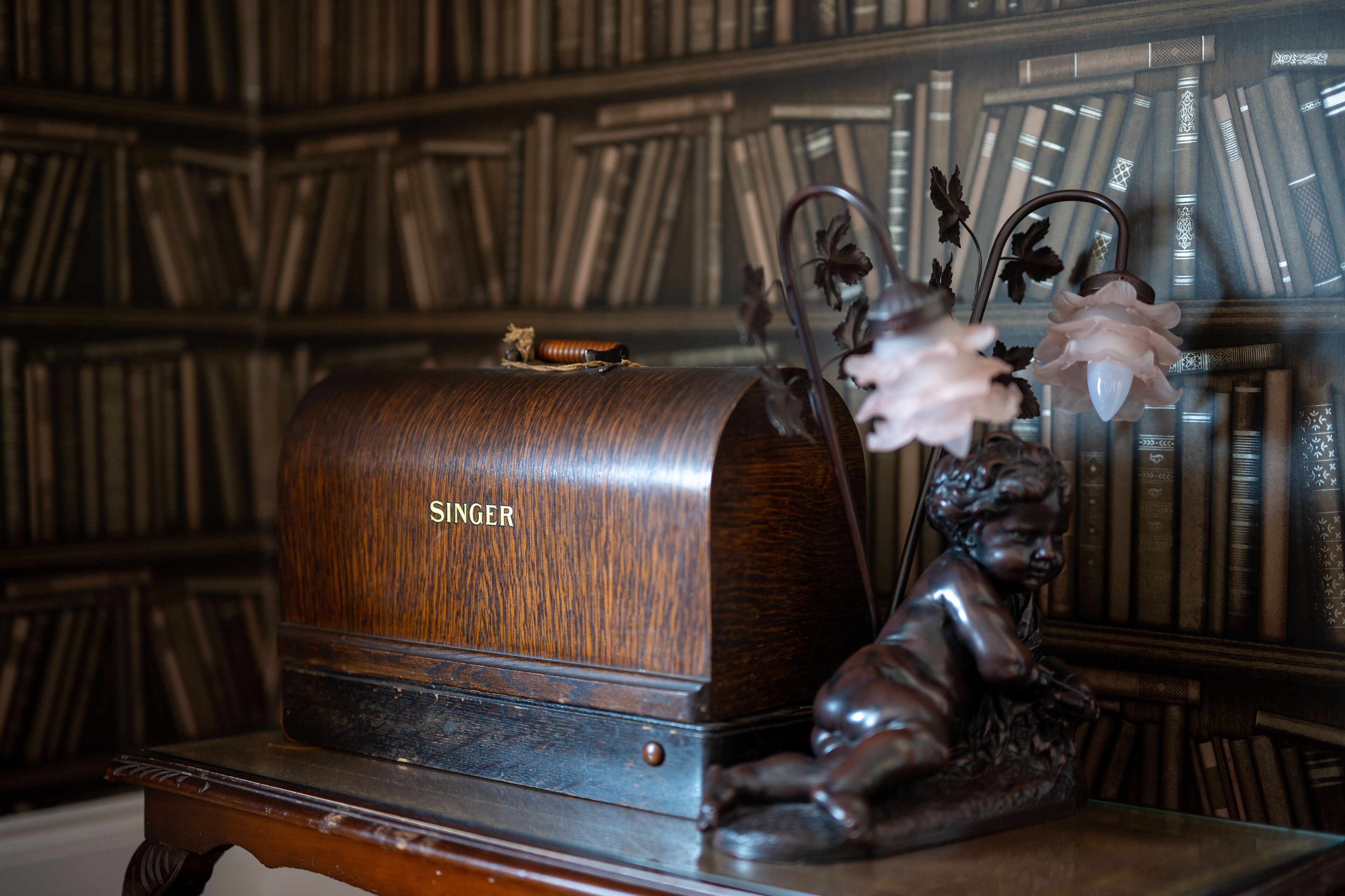 Una máquina de escribir Singer de madera sobre una mesa, junto a una figurita de ángel de latón, con una estantería llena de libros como fondo.