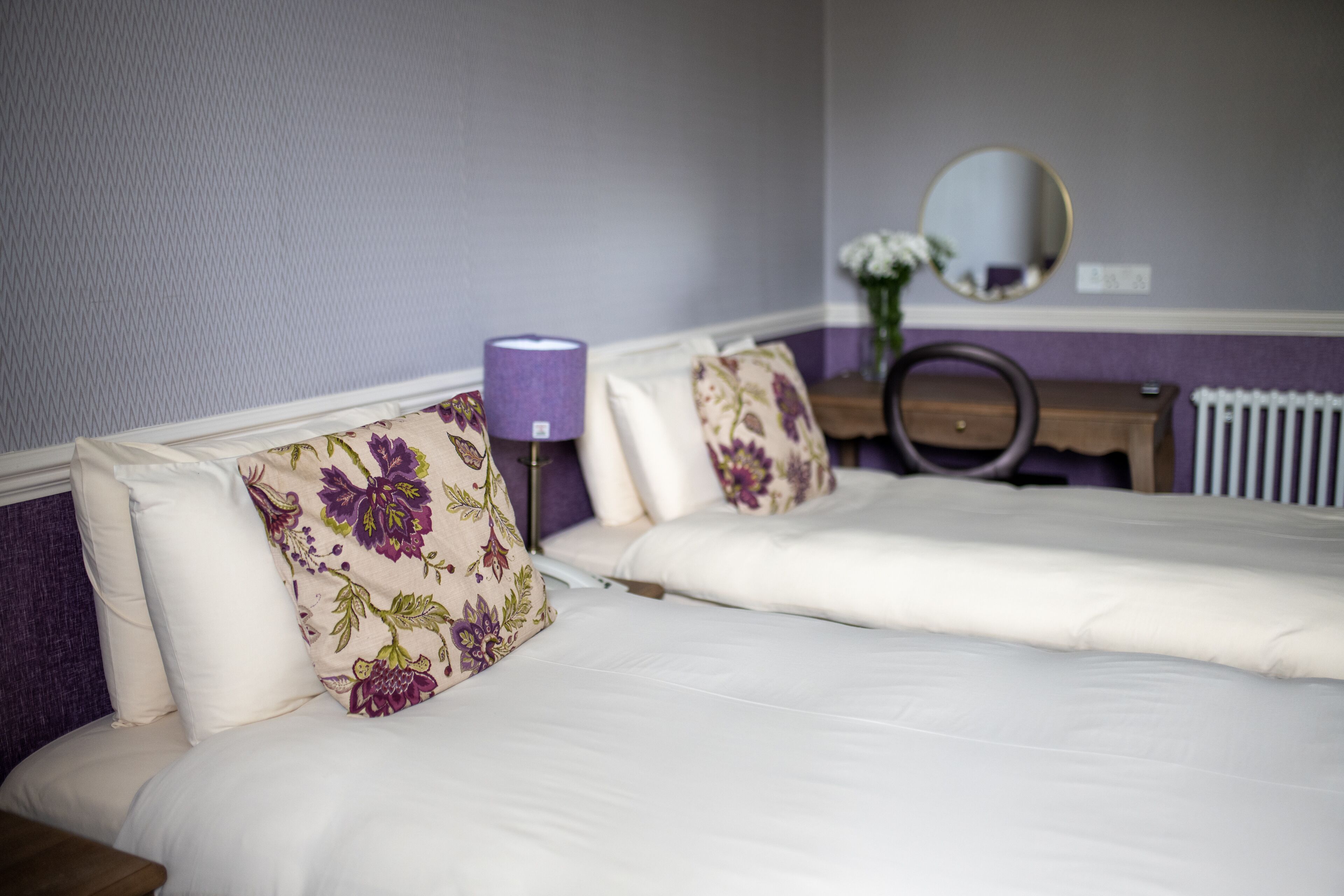 Ein Doppelbett mit blumigen Kissen, ein Nachttisch mit Lampe und ein Spiegel an der Wand.