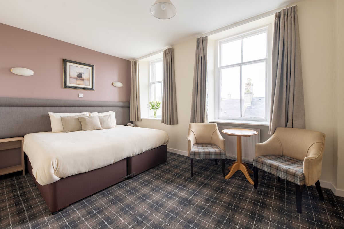 A hotel room with a bed, a chair, a small table, and a window with curtains.