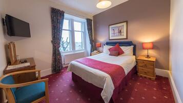 A hotel room with a bed, a desk with a chair, a TV mounted on the wall, and a window with curtains.
