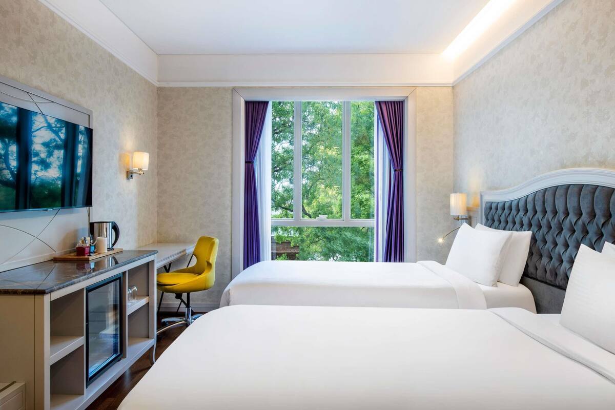 Une chambre d’hôtel avec un lit, un bureau et une chaise, une télévision et une fenêtre avec des rideaux.
