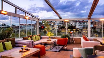 Un moderno bar en la azotea con vistas panorámicas de la ciudad, amueblado con sofás, mesas y sillas.