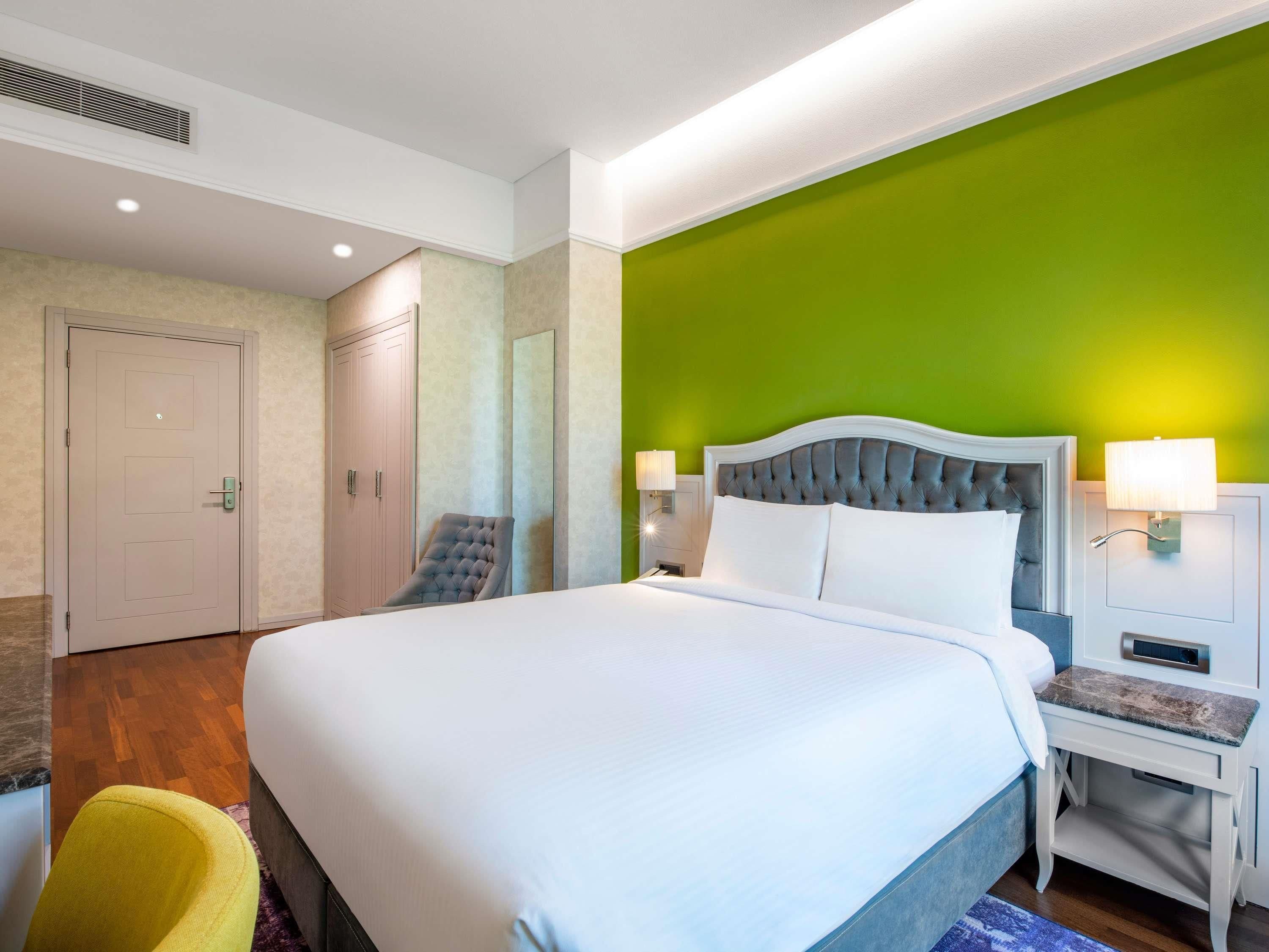 A hotel room with a large bed, a chair, a desk, and a green accent wall.