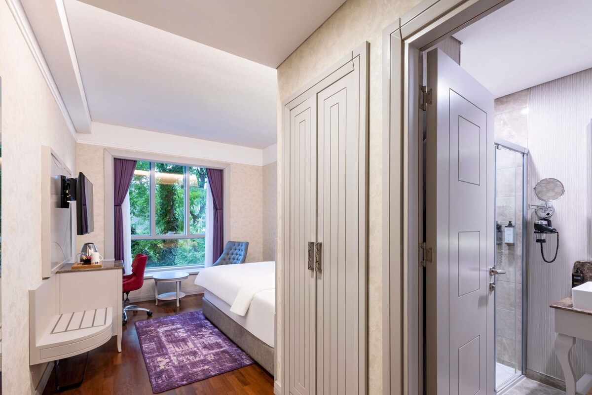 Habitación de hotel con cama, escritorio con silla, baño con ducha y ventana con cortinas.