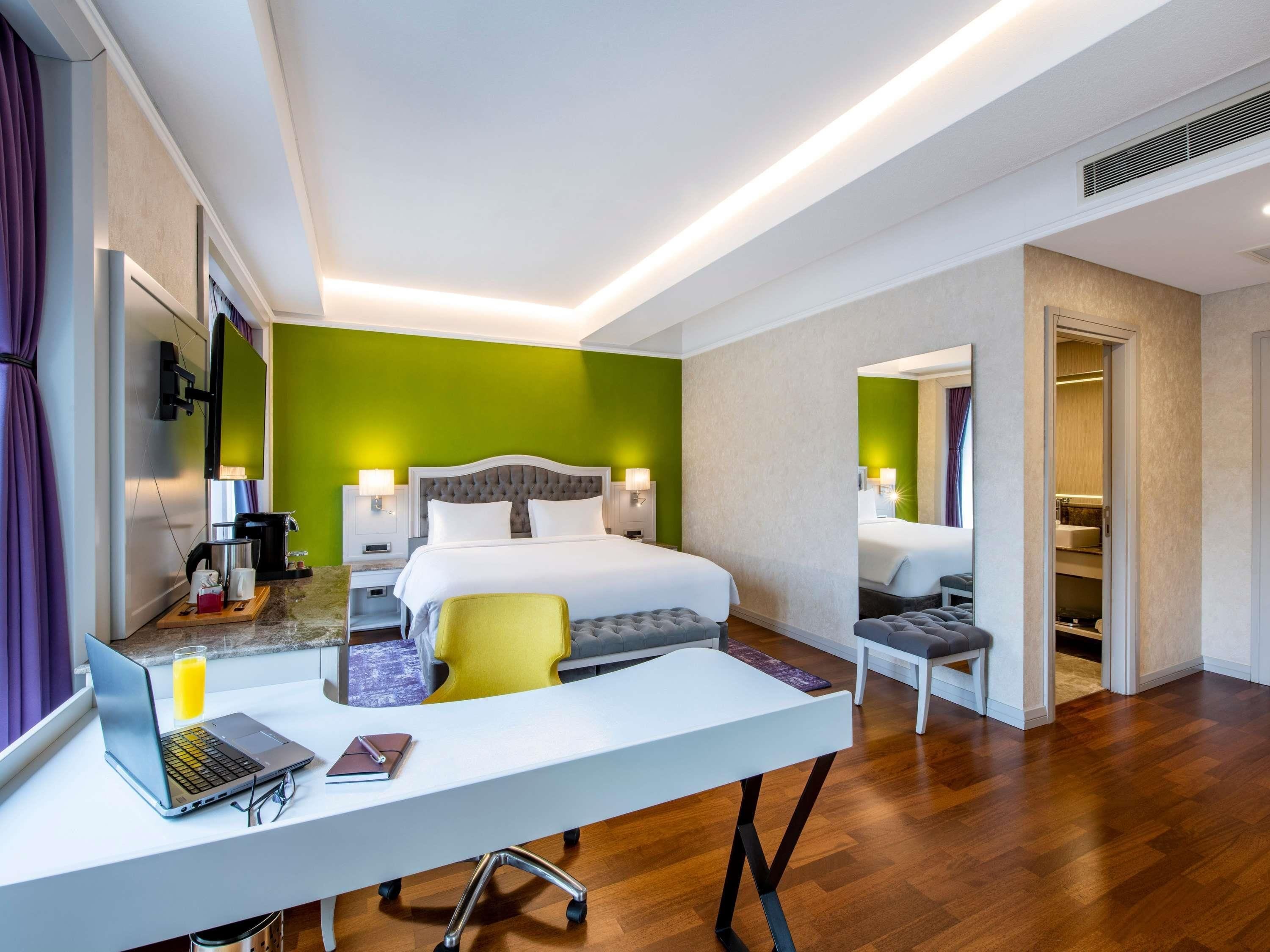A modern hotel room with a large bed, a desk with a laptop, a chair, and a small table.