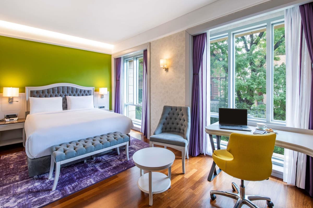 Une chambre d’hôtel avec un lit, un bureau, une chaise et une vue sur les arbres, visible à travers de grandes fenêtres.
