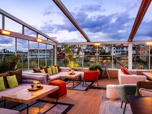 A modern rooftop lounge with large windows, a sofa set, a dining table, and a red chair.