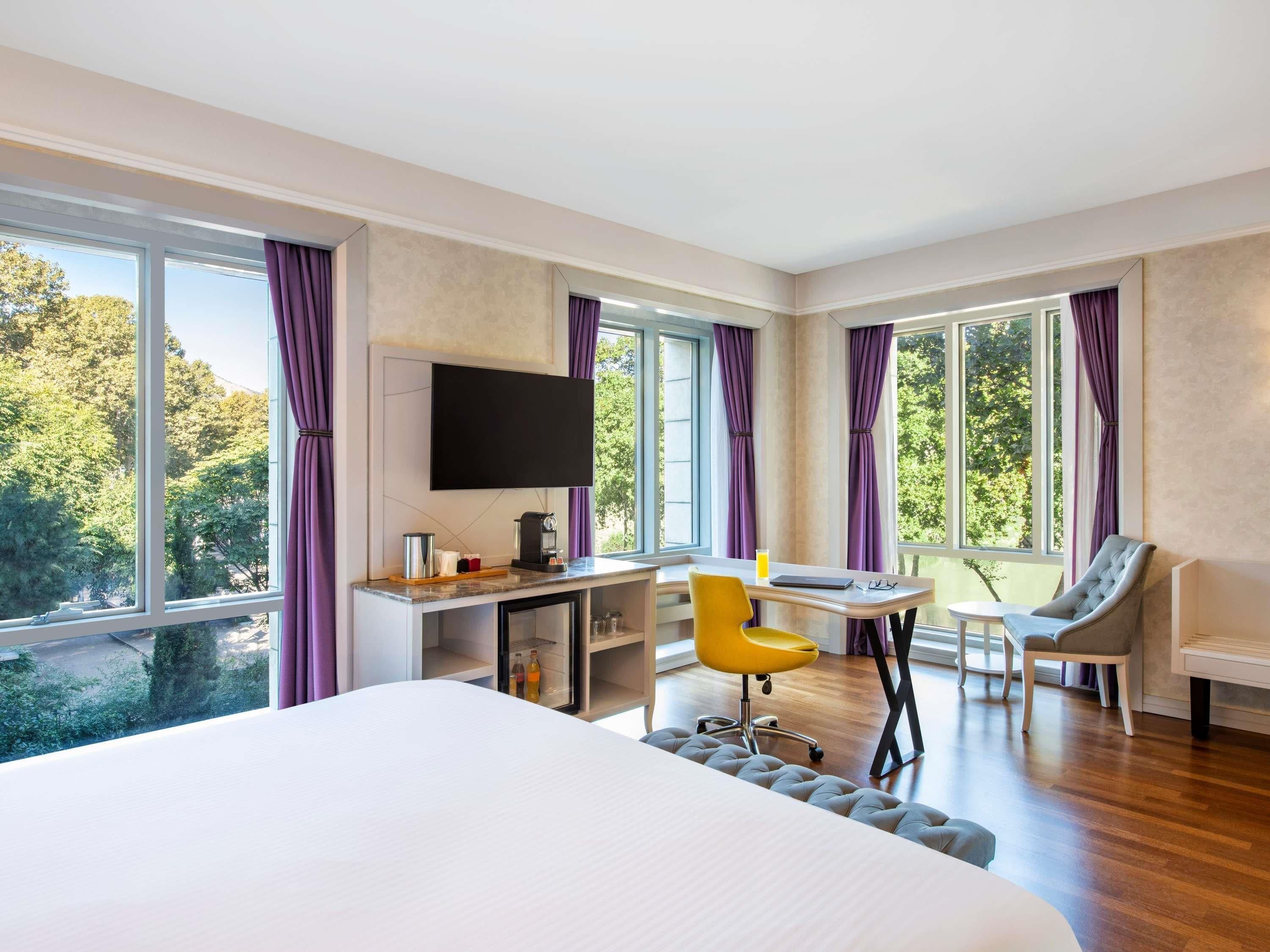 Una habitación de hotel moderna con una cama grande, un escritorio con televisor de pantalla plana, una silla amarilla y ventanas amplias con cortinas.