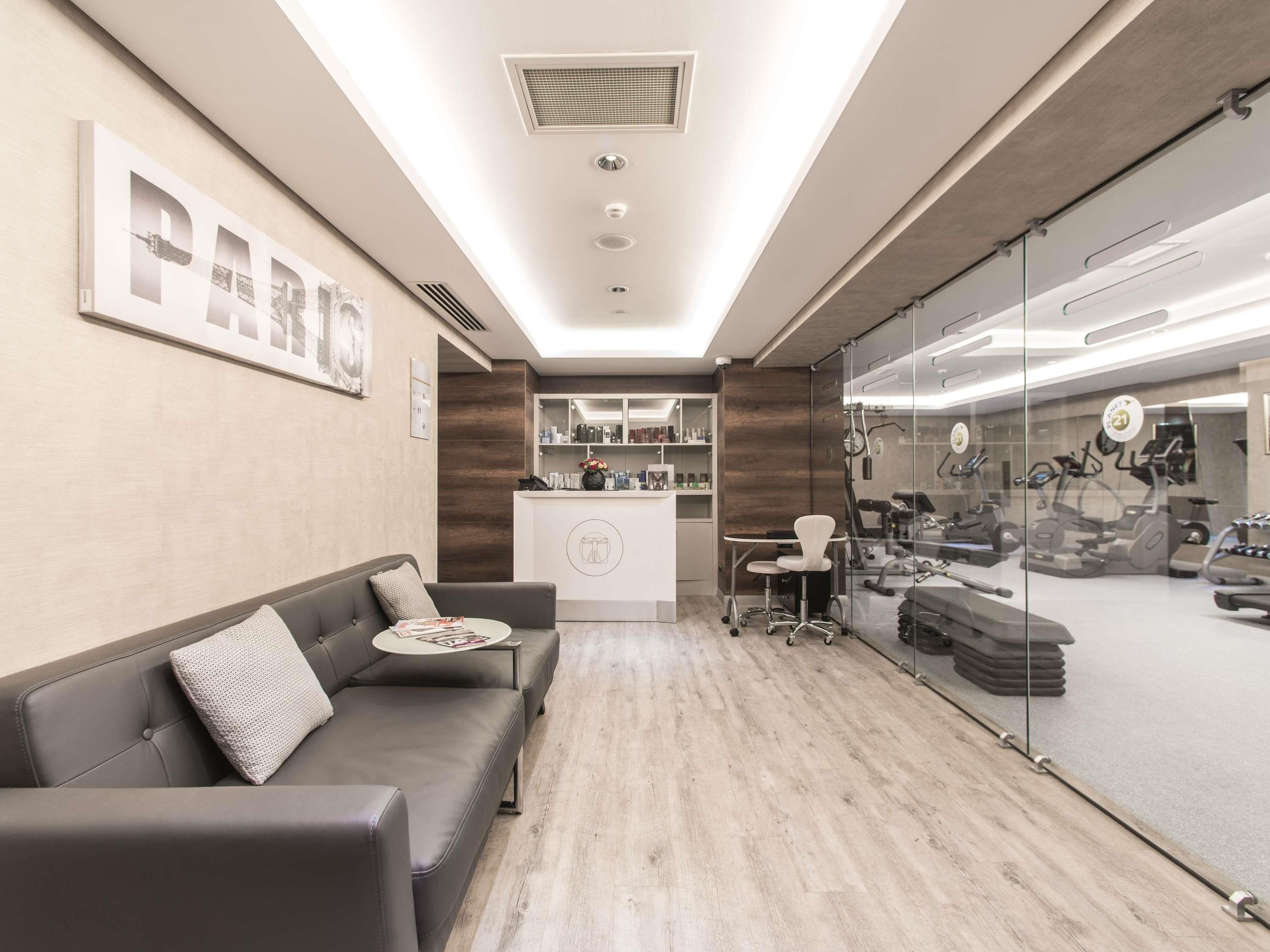 Een receptiezone met een bank, een receptiebalie, een glazen wand met fitnessapparatuur en een houten vloer.