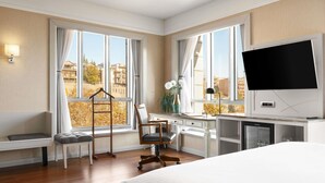 Habitación de hotel con cama, escritorio, silla y ventanas amplias que dan a vistas de edificios y árboles.