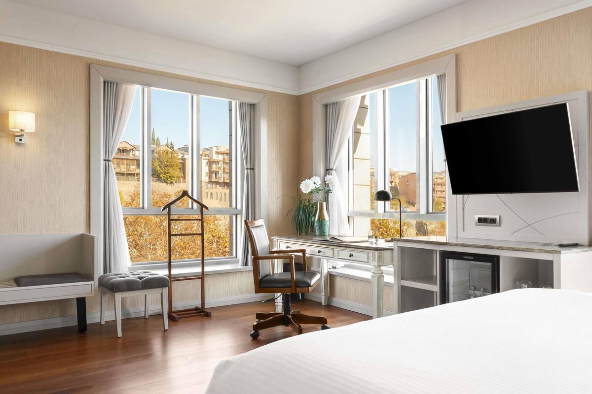 A hotel room with a bed, desk, chair, and large windows offering a view of buildings and trees.