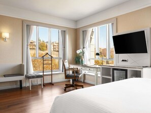 Ett hotellrum med en säng, ett skrivbord, en stol och stora fönster med utsikt över byggnader och träd.