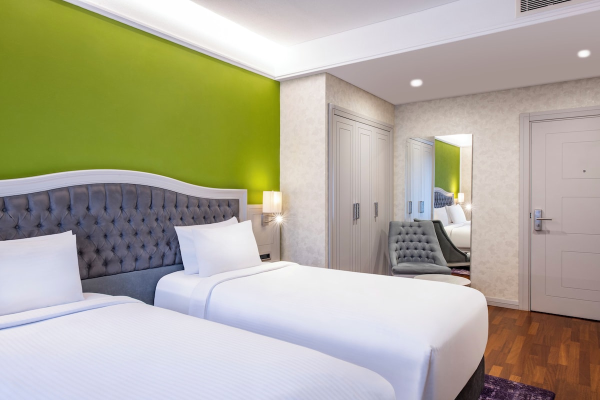 Habitación de hotel con dos camas, una pared de acento verde, un cabecero capitonado, un sillón gris y un espejo que refleja la habitación.