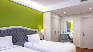 A hotel room with two beds, a green accent wall, a tufted headboard, a grey armchair, and a mirror reflecting the room.
