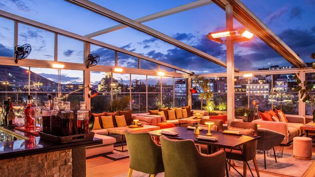 Un bar en la azotea con mesas, una barra con botellas y vistas a la ciudad.