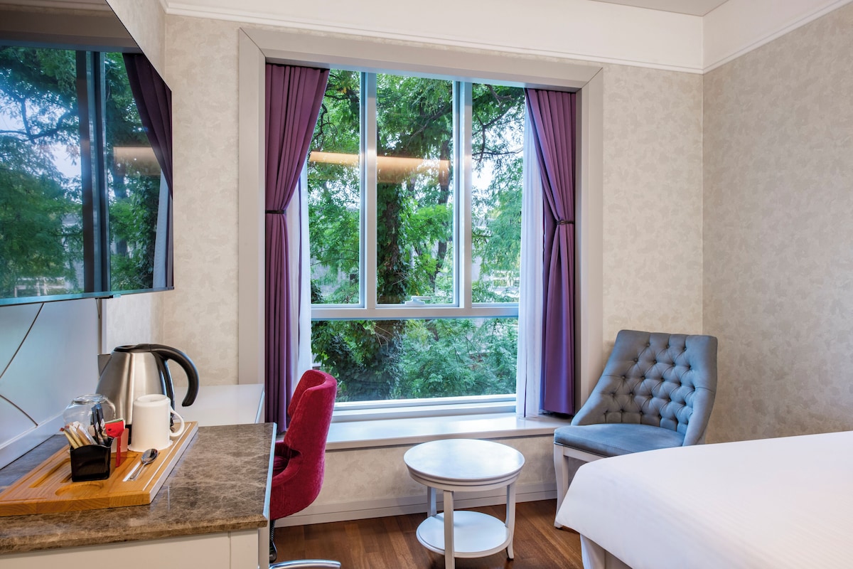 Habitación de hotel con cama, una mesita, una silla, ventana con cortinas y vista a árboles.