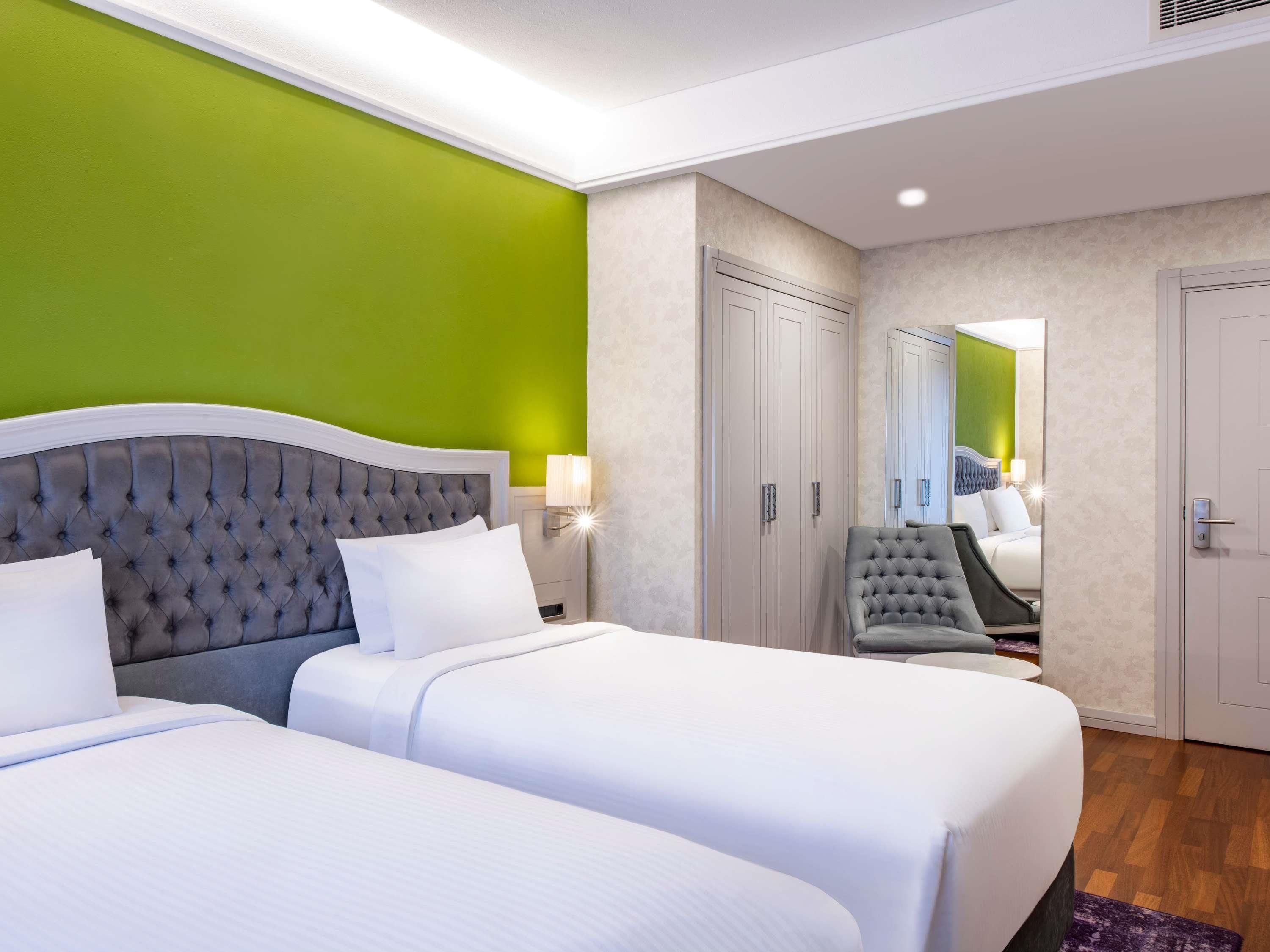 Ein Hotelzimmer mit zwei Betten, einer grünen Akzentwand, einem großen Spiegel und einem Kleiderschrank.