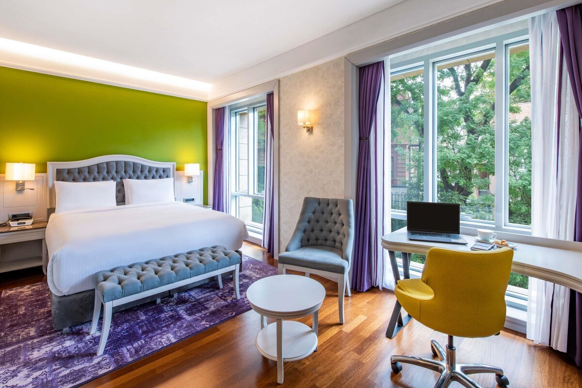 Habitación de hotel con cama, escritorio, silla y vistas a árboles a través de ventanas amplias.