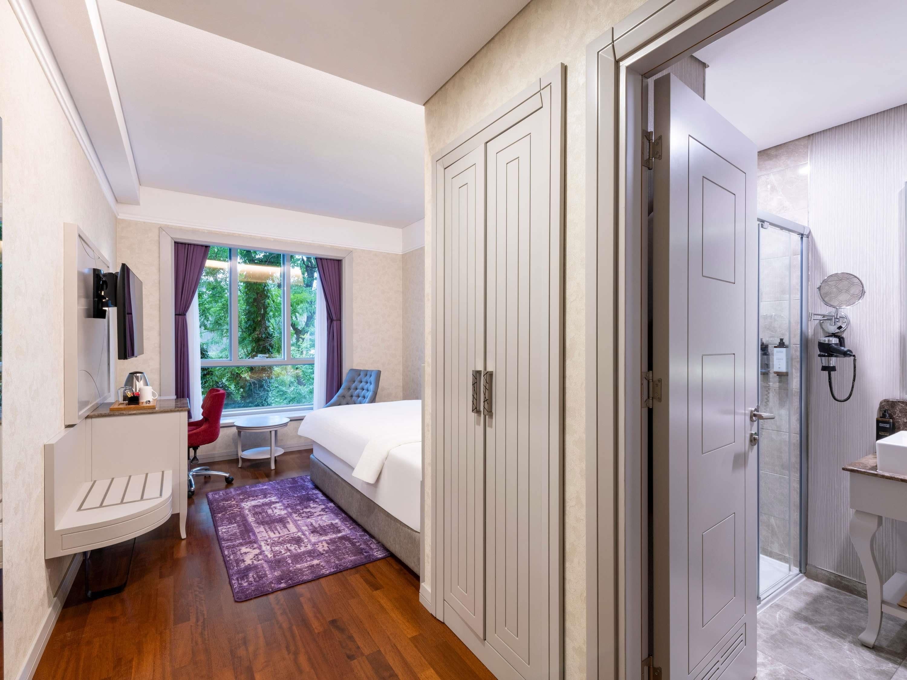 Quarto de hotel com cama, escrivaninha com cadeira, banheiro com chuveiro e janela com cortinas.