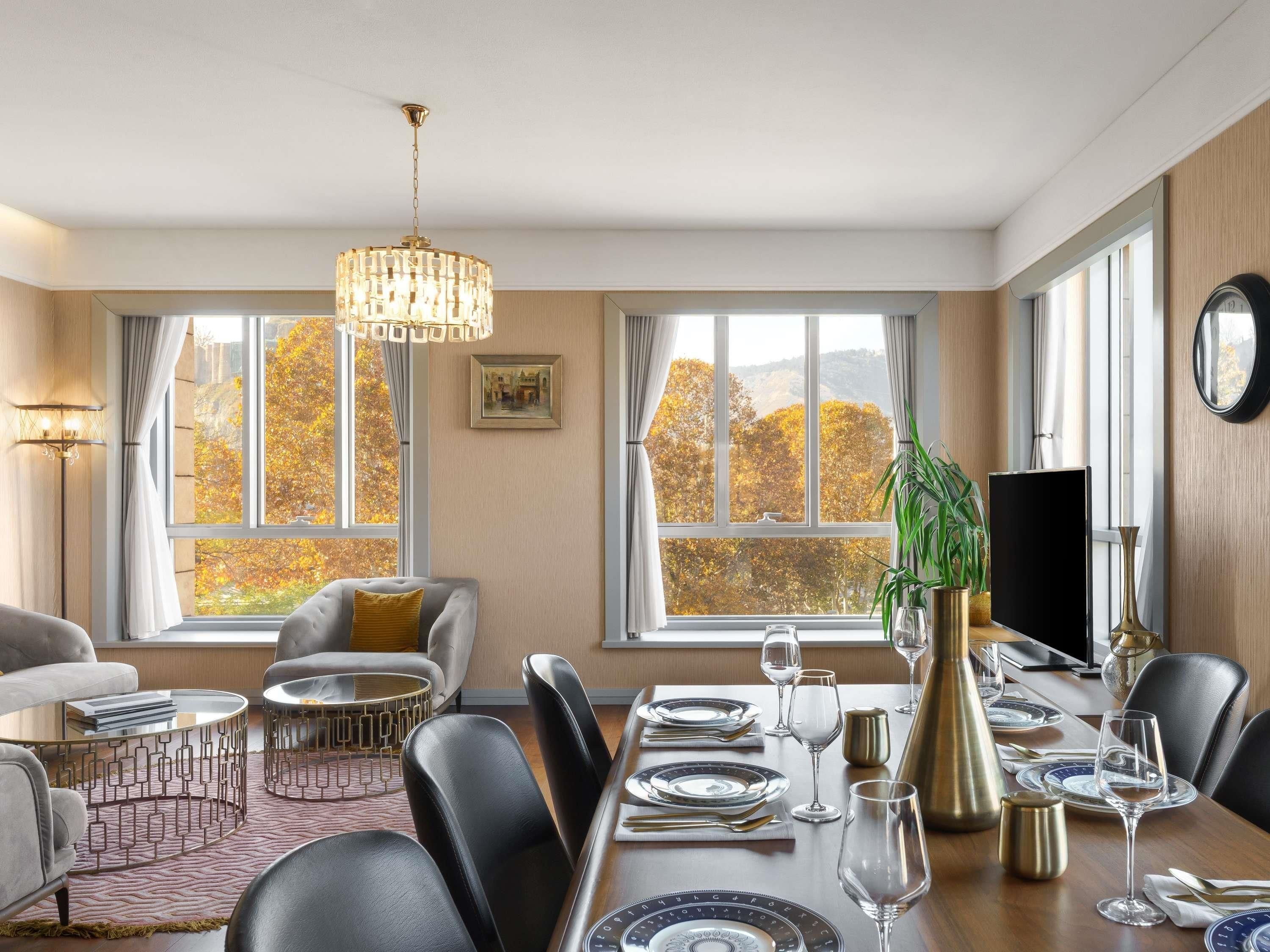Een eethoek met een tafel voor vier personen, een bank en een groot raam met uitzicht op herfstbladeren.