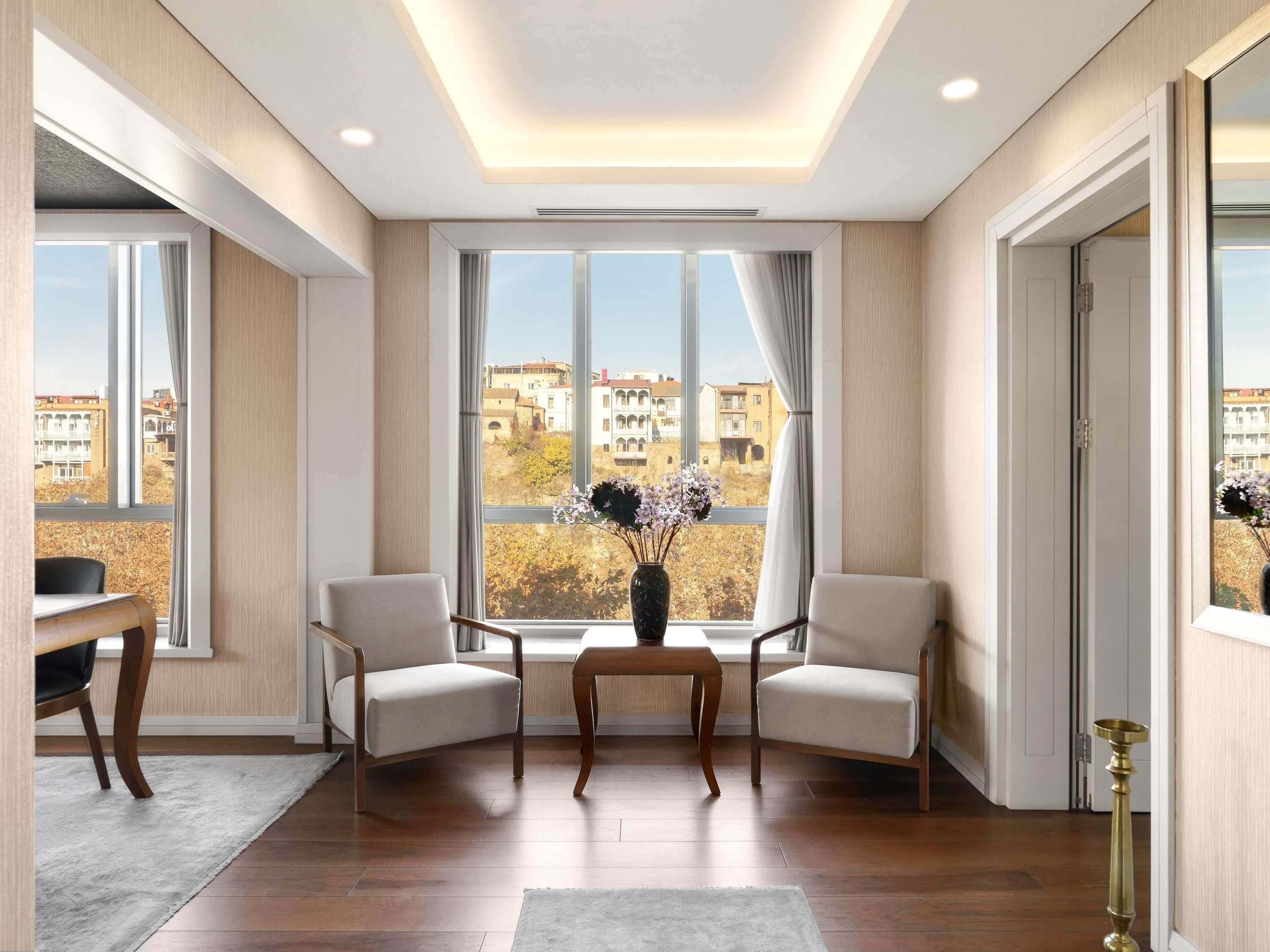 Un salon moderne avec une grande fenêtre, deux fauteuils, une petite table et un vase rempli de fleurs.