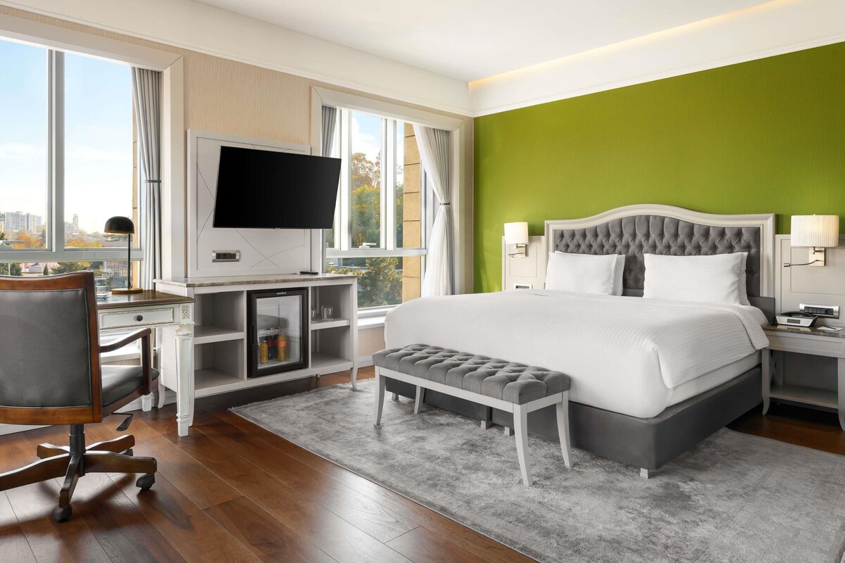 A hotel room with a bed, a desk with a television, a small refrigerator, a bench, and a view of the city through large windows.