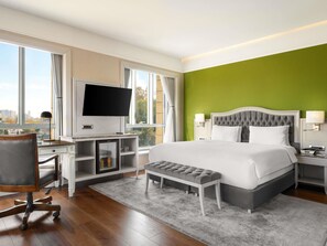 Habitación de hotel con cama, escritorio con televisor, refrigerador pequeño, banco y vista de la ciudad a través de amplias ventanas.