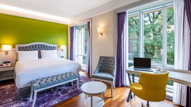 A hotel room with a bed, a desk, a chair, and a view of trees through large windows.