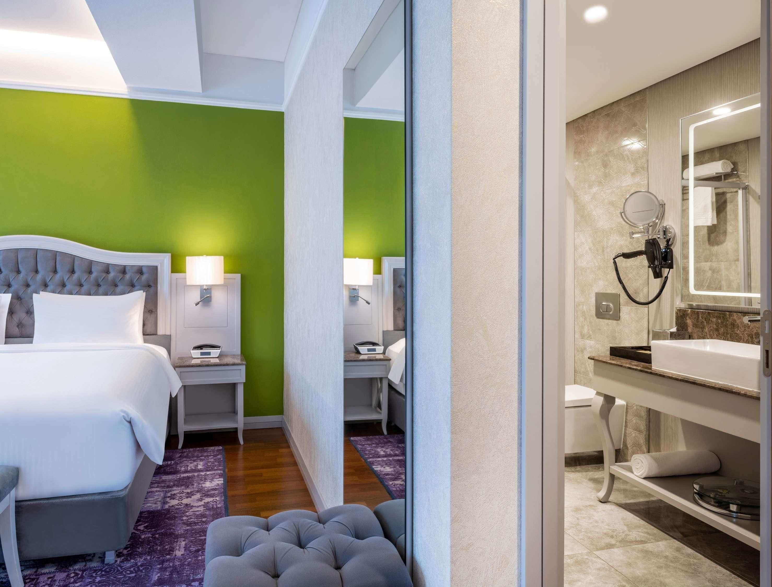 A hotel room with a bed, bedside tables, a mirror, a bathroom with a sink and mirror, and a purple rug.