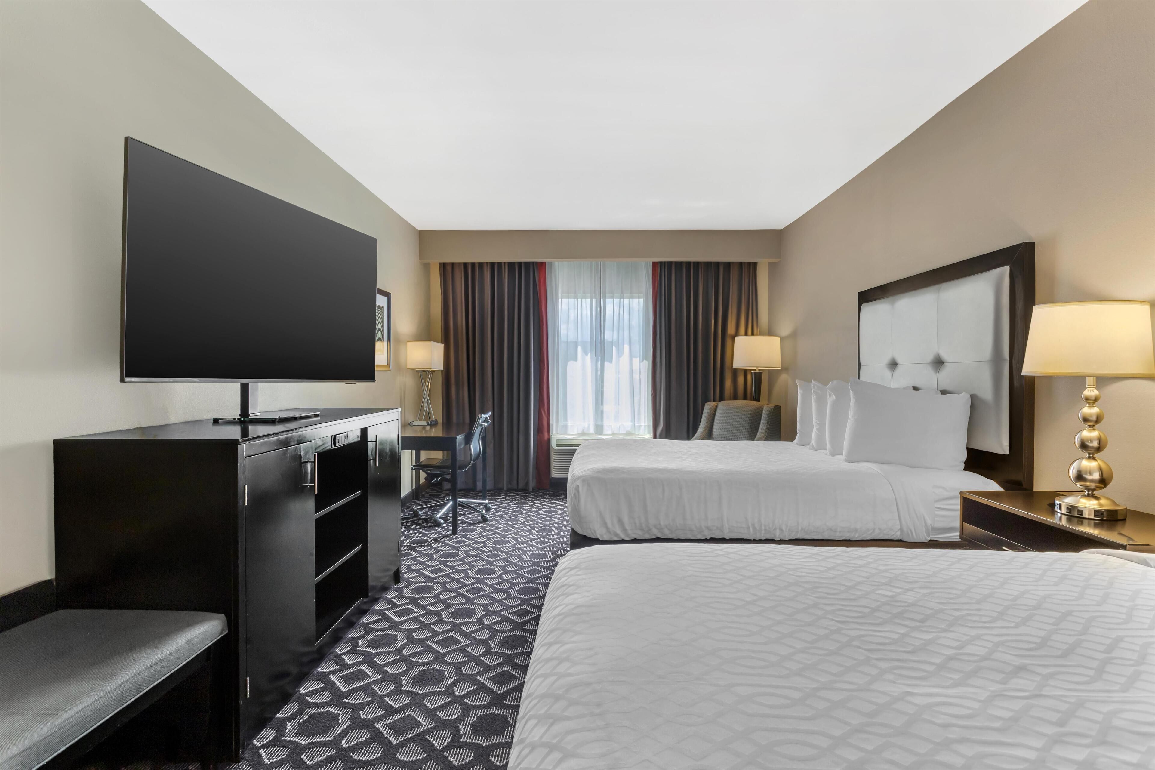 A hotel room with two beds, a desk with a chair, a flat-screen TV, and a large window with curtains.