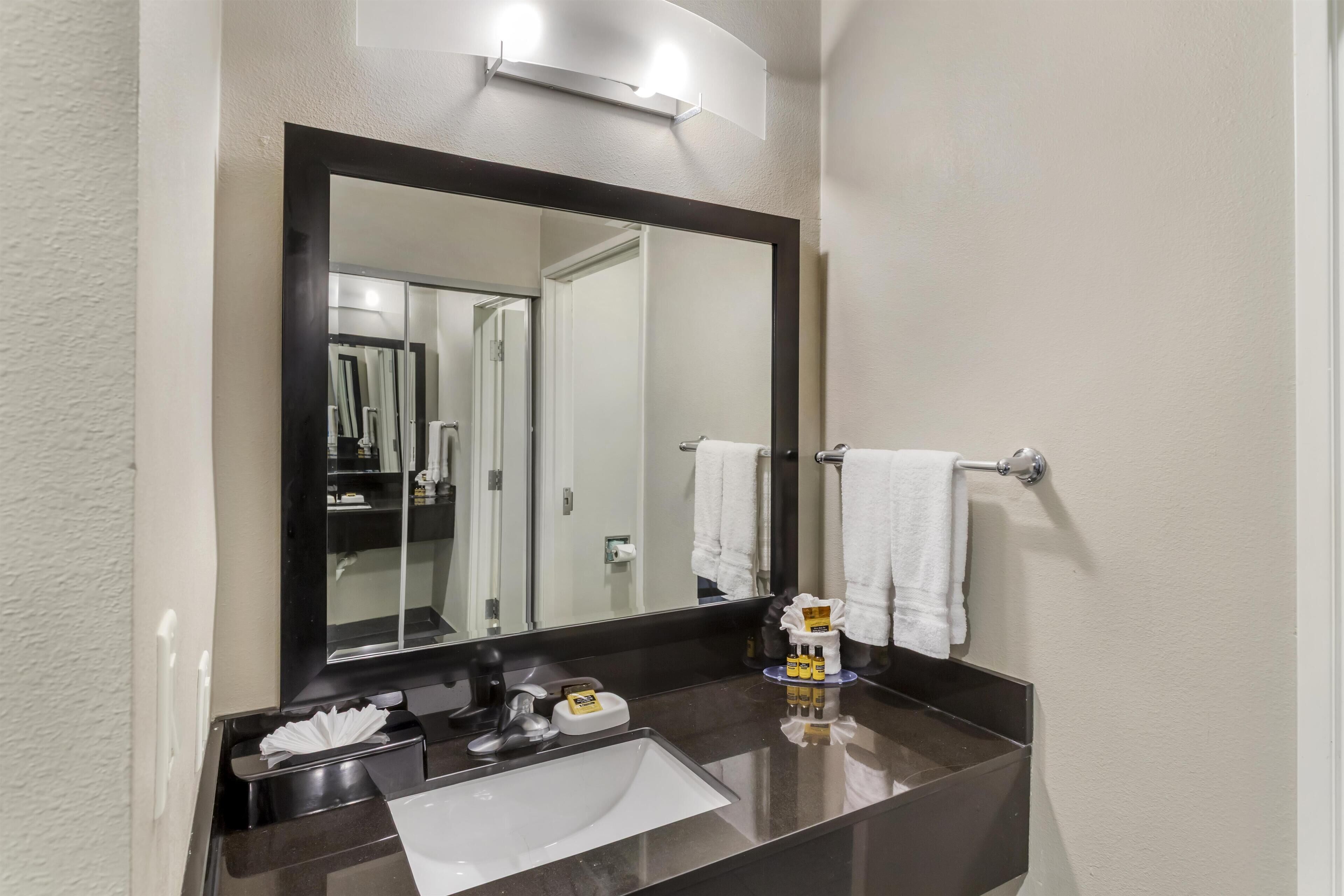 A bathroom with a dark countertop, a white sink, a large mirror, and a towel rack with white towels.