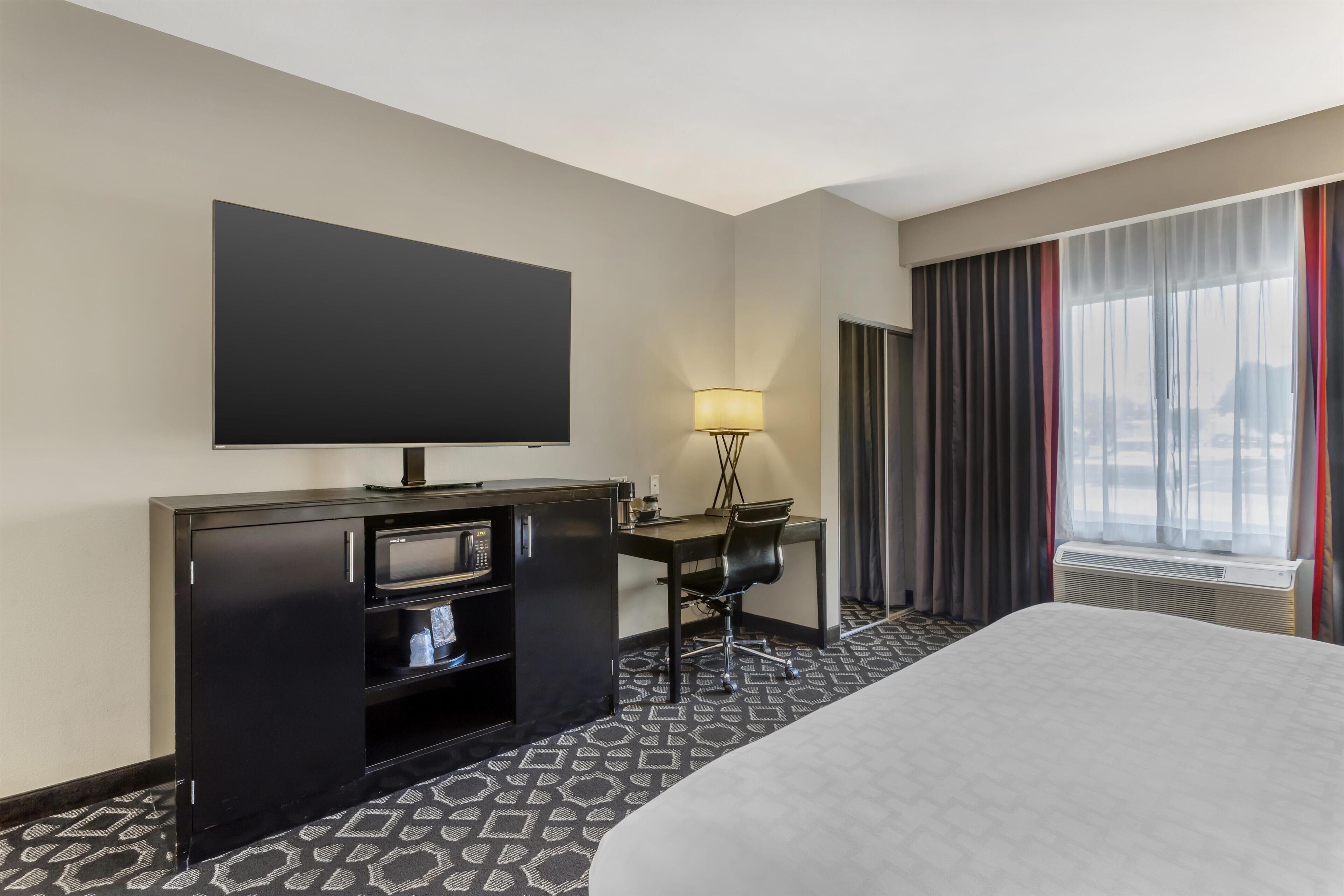 Ein Hotelzimmer mit einem großen Bett, einem flachbildfernseher an der Wand, einem Schreibtisch mit Stuhl, einer Lampe und einem fenster mit Vorhängen.