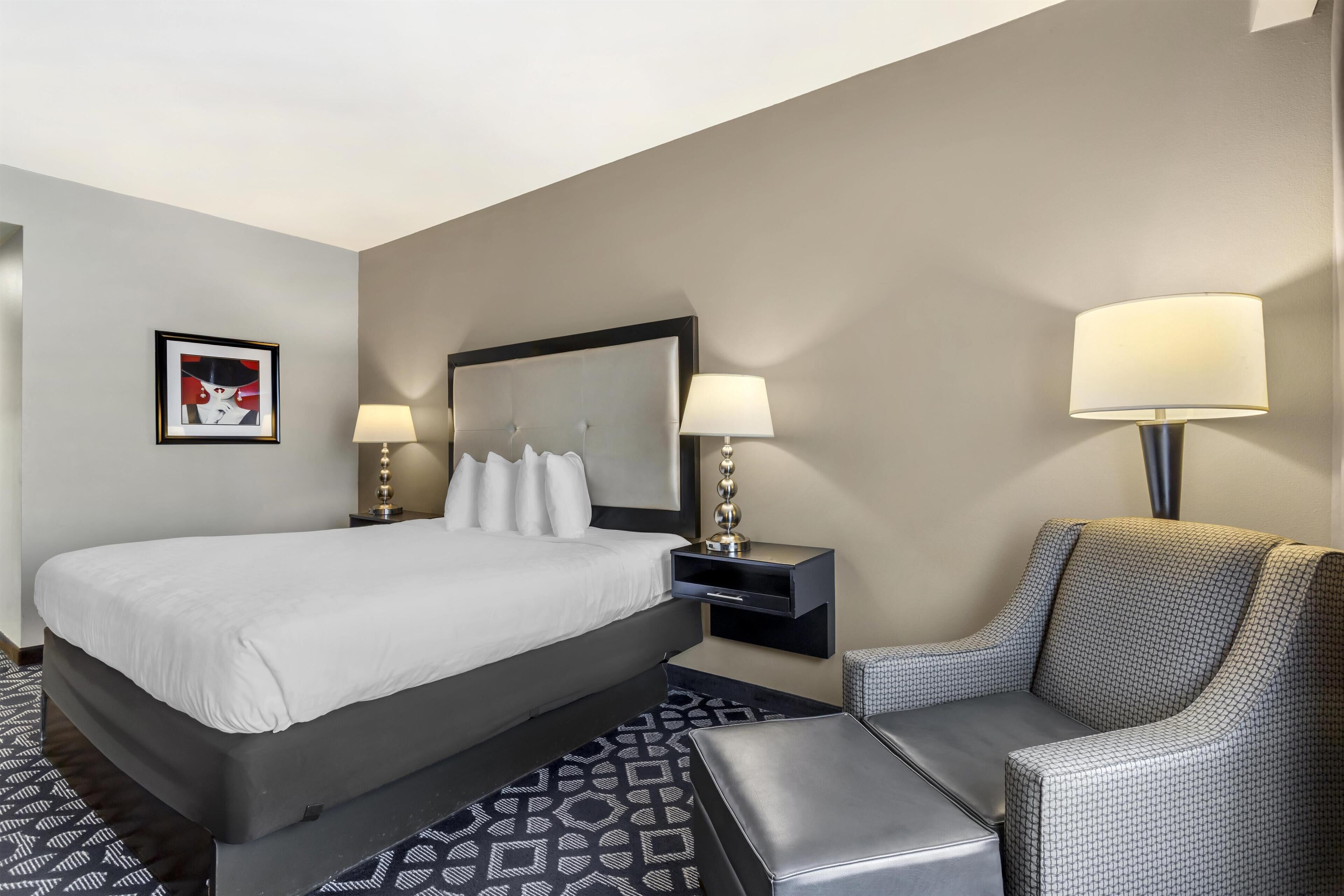 A hotel room with a large bed, two bedside tables with lamps, a chair, a patterned rug, and a framed artwork on the wall.