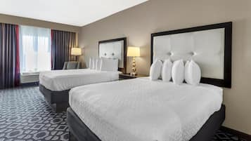 Quarto de hotel com duas camas, uma janela grande com cortinas, uma TV fixada na parede e uma mesa de cabeceira com abajur.