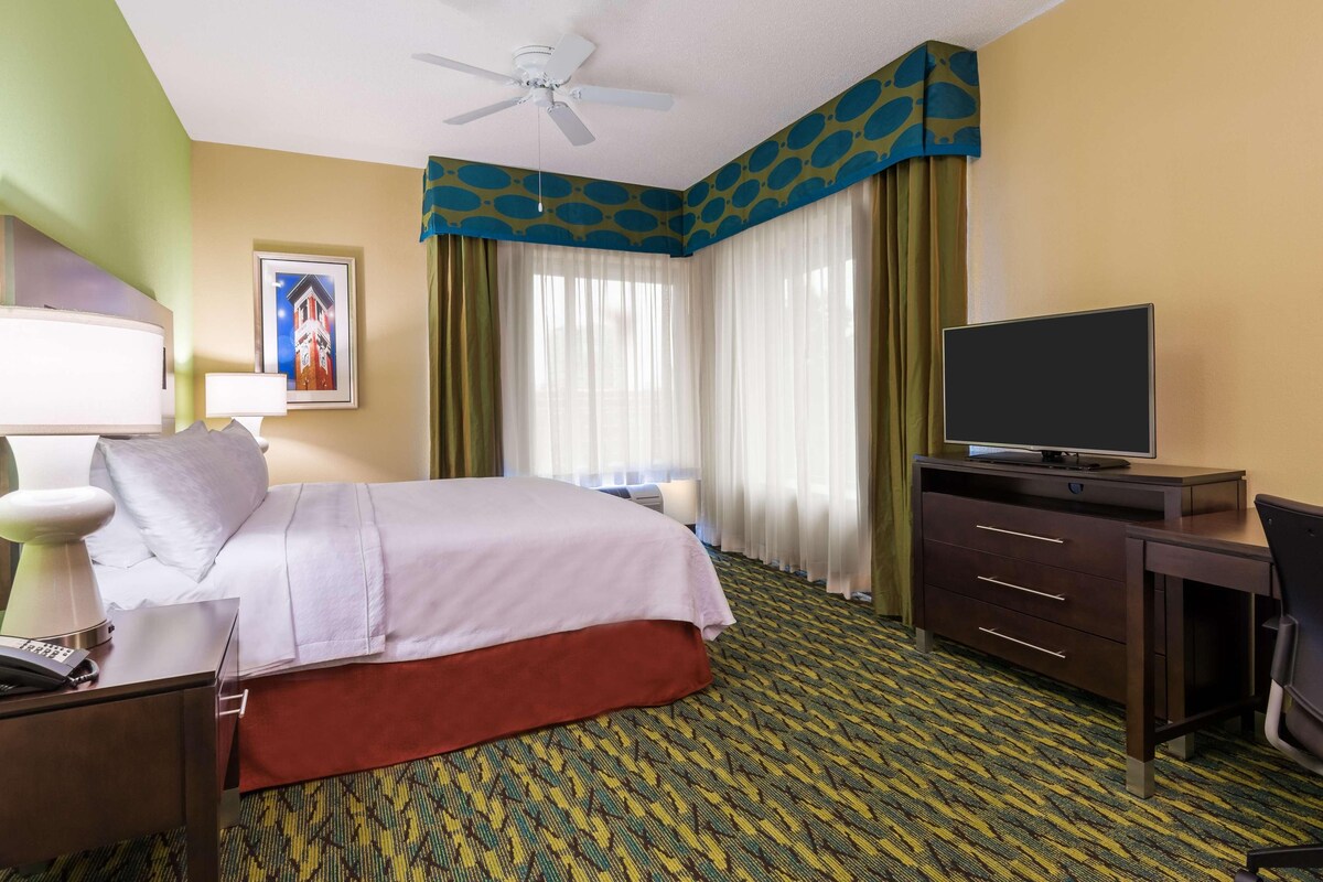 Habitación de hotel con cama, escritorio, televisor, ventilador de techo y alfombra estampada.