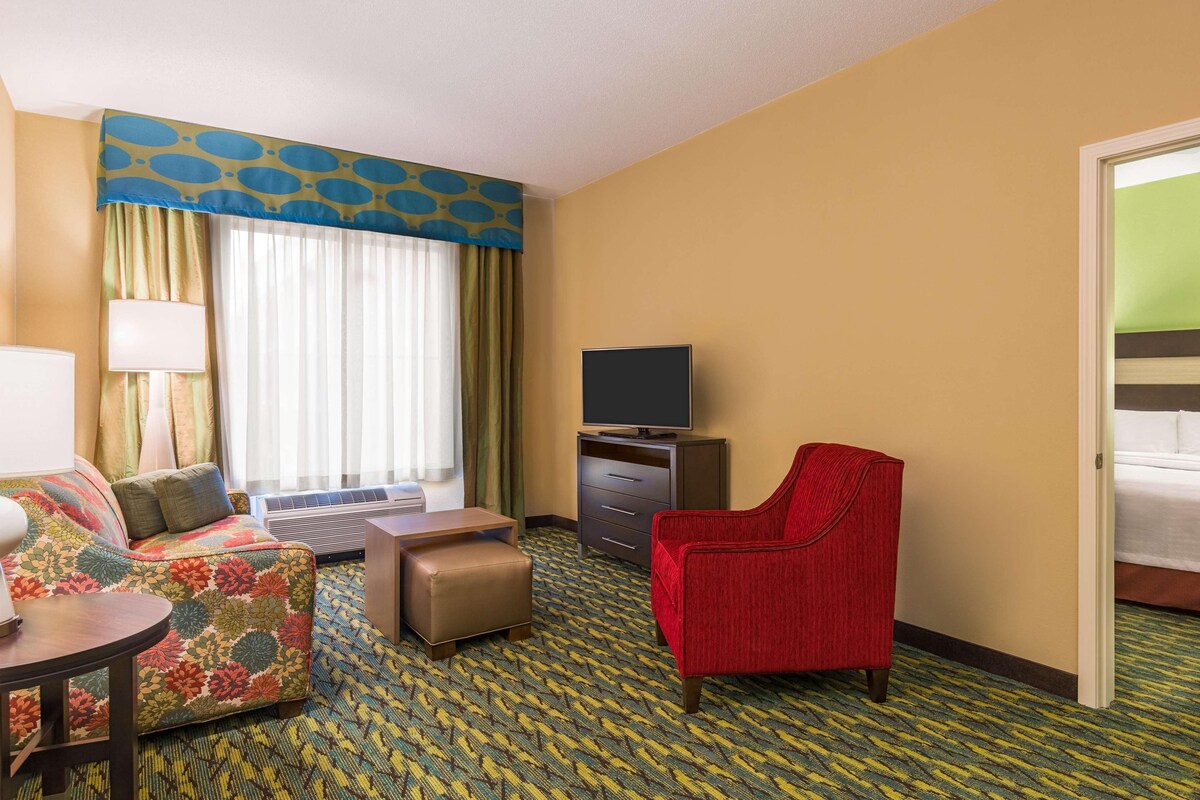 Habitación de hotel con sofá, sillón rojo, televisor en un mueble bajo, una mesita y una ventana con cortinas.