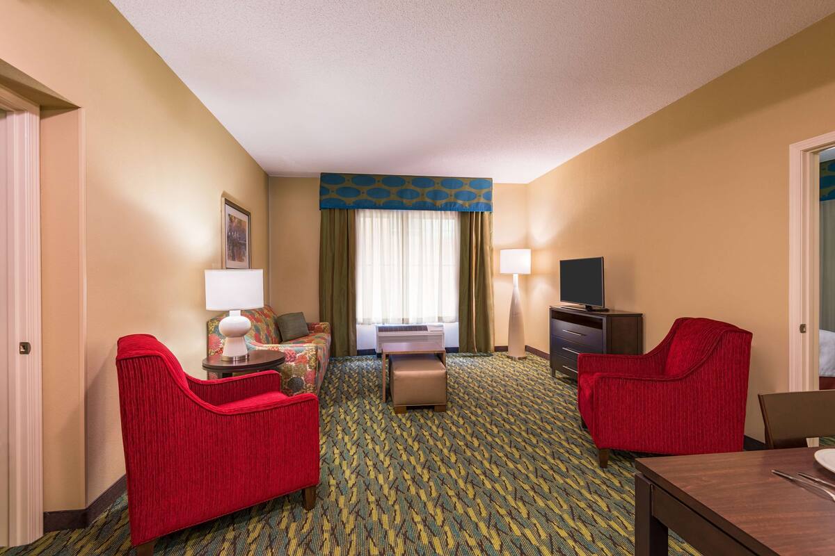 A hotel room with a bed, two red armchairs, a small table with a lamp, a TV, and a carpeted floor.