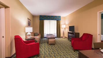 A hotel room with a bed, two red armchairs, a small table with a lamp, a TV, and a carpeted floor.