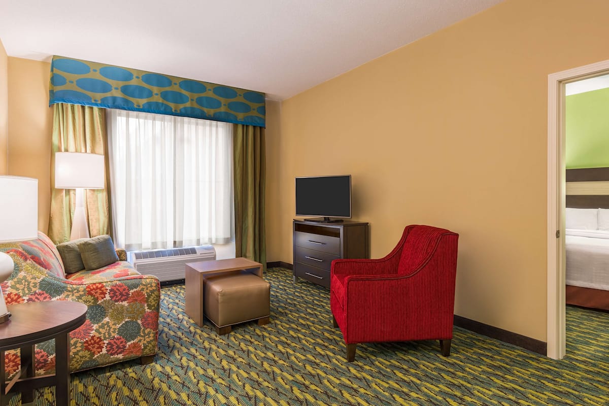 A hotel room with a sofa, a red armchair, a television, a desk, and a window with curtains.