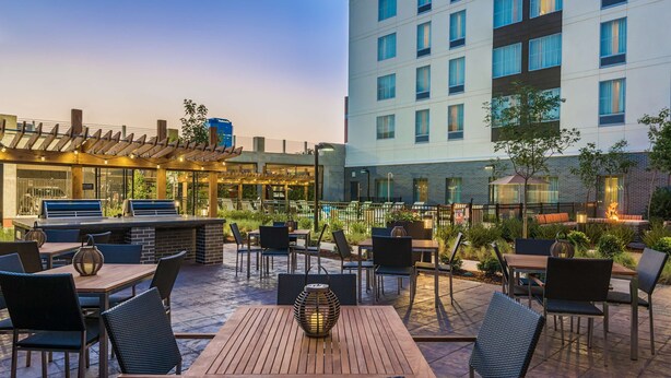 An outdoor patio with wooden tables and chairs, a bar area, and a view of a modern building.