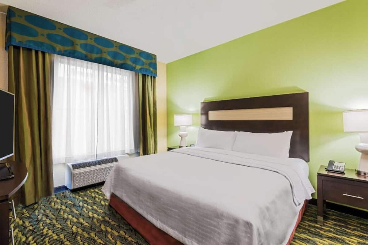Habitación de hotel con cama, escritorio, silla, televisión y ventana con cortinas.