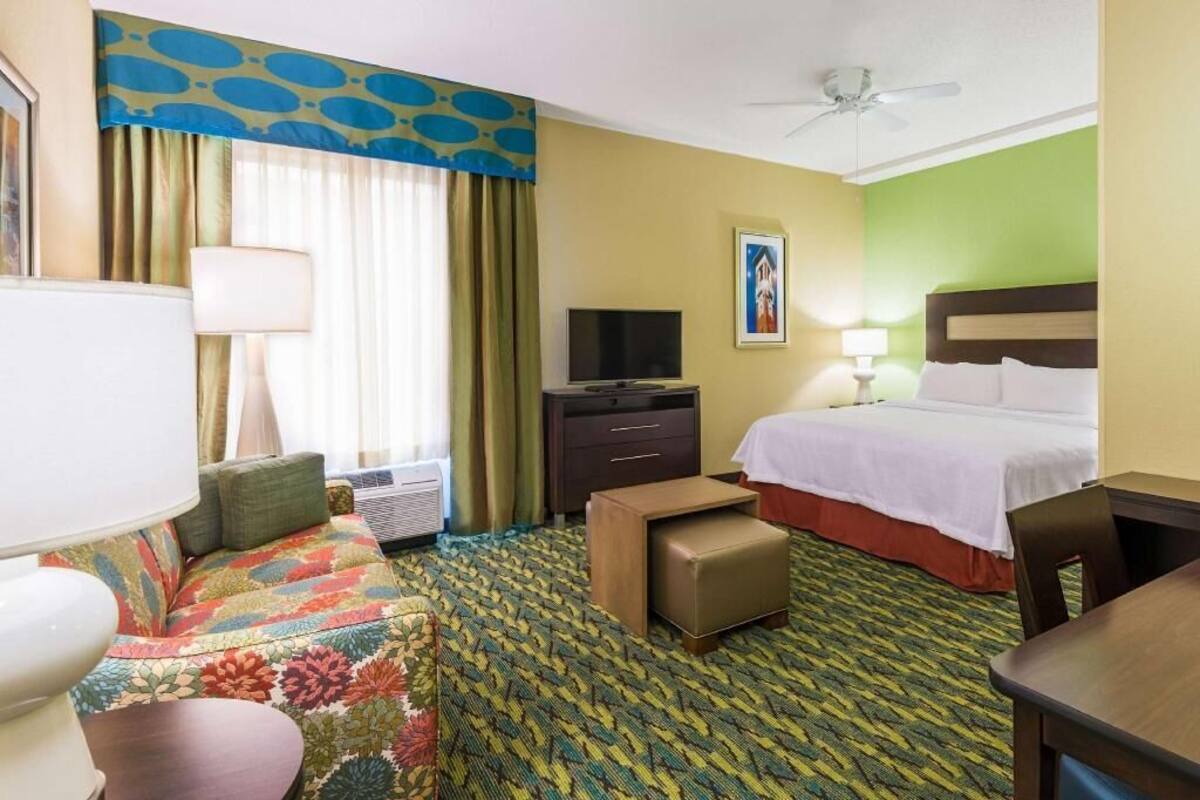 A hotel room with a bed, a TV, a desk, a chair, a sofa, a lamp, and a ceiling fan.
