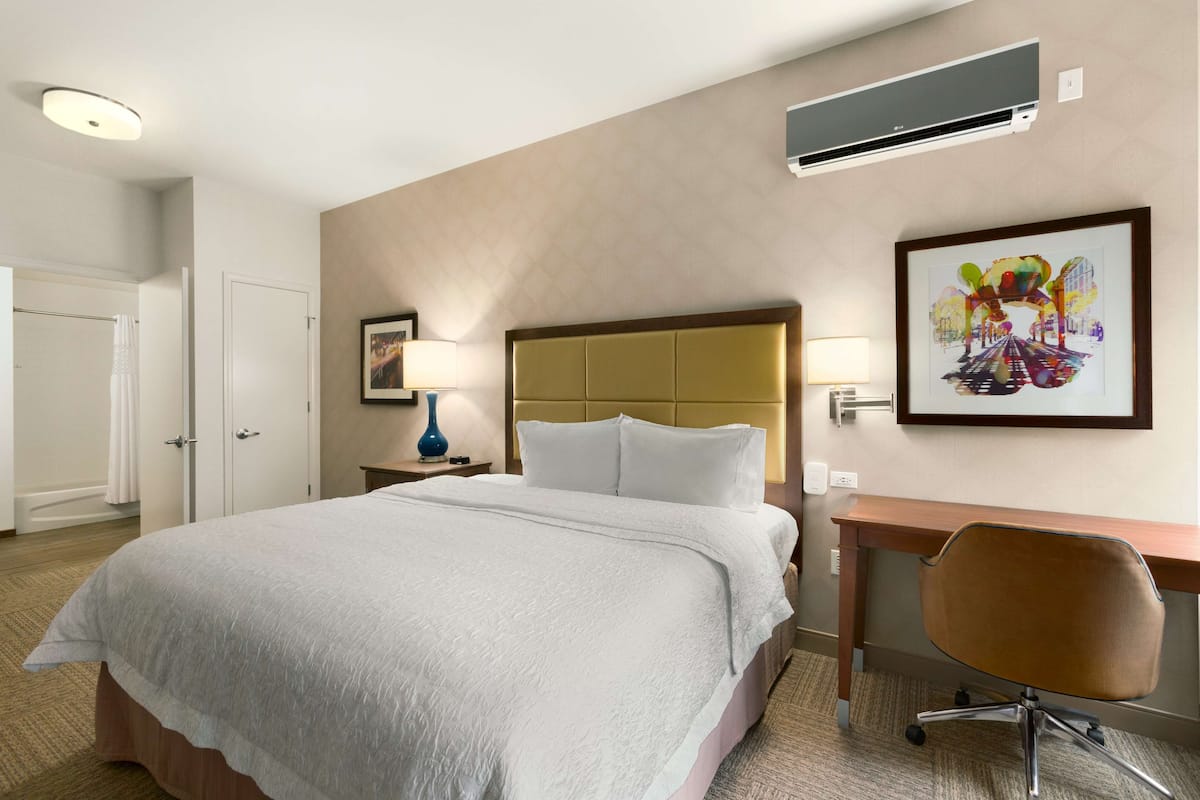 A hotel room with a large bed, a desk, a chair, an air conditioner, and a framed picture on the wall.
