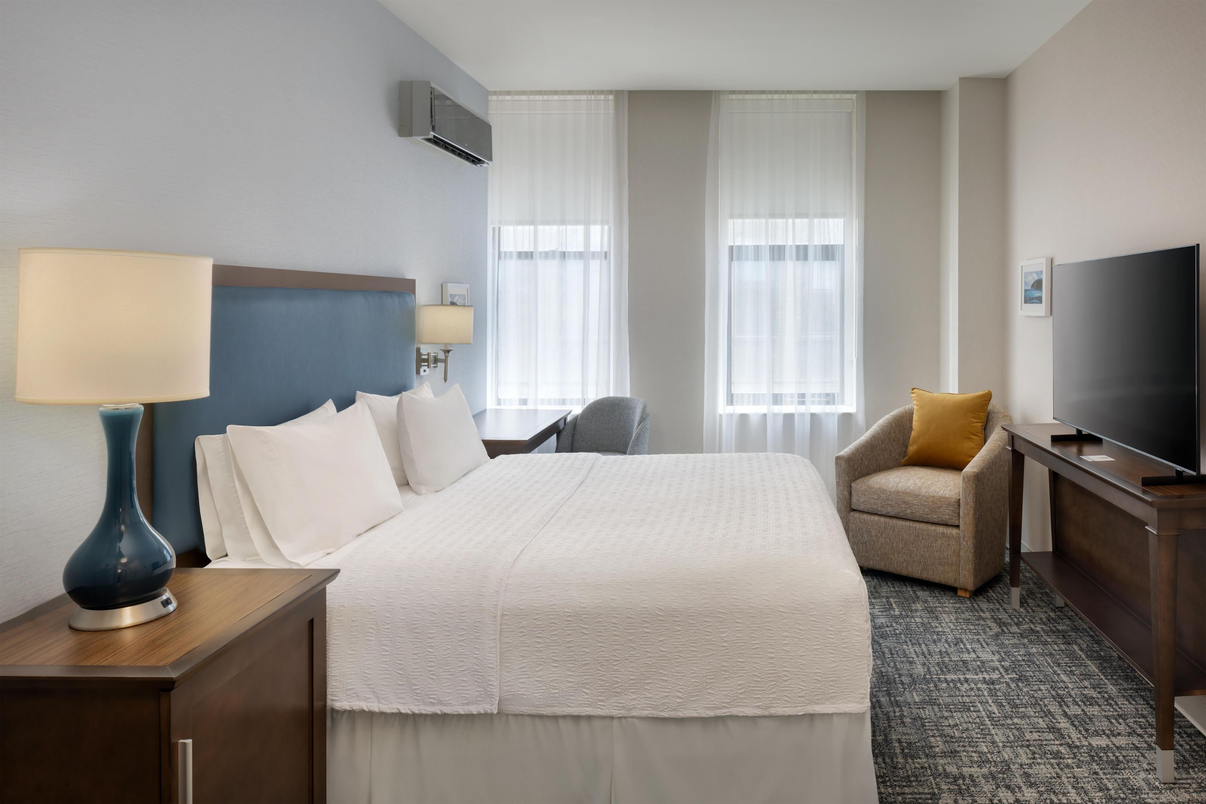 Una habitación de hotel con una cama, un escritorio con lámpara, una silla, un televisor y dos ventanas con cortinas.