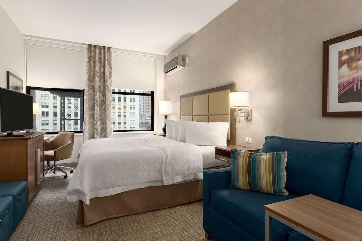 A hotel room with a large bed, a blue sofa, a desk with a TV, and a window with a city view.