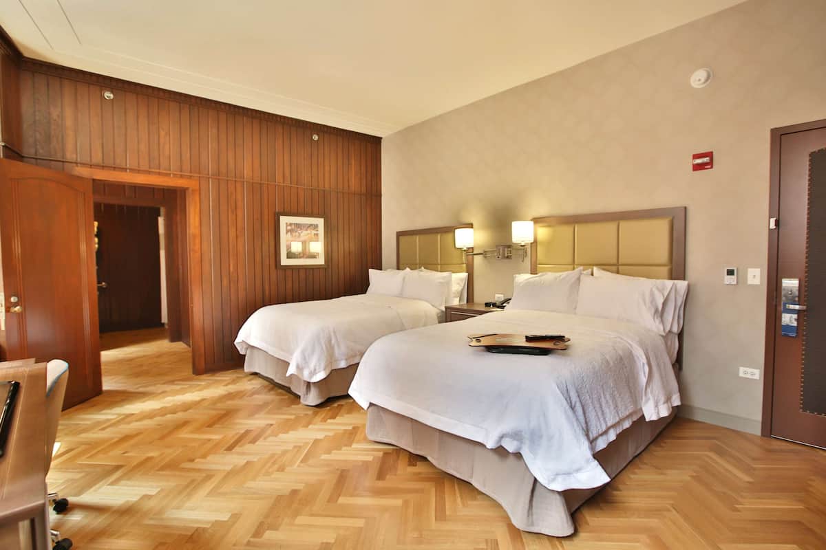 A hotel room with two beds, wooden paneling on the walls, a wooden herringbone floor, a desk, and a chair.