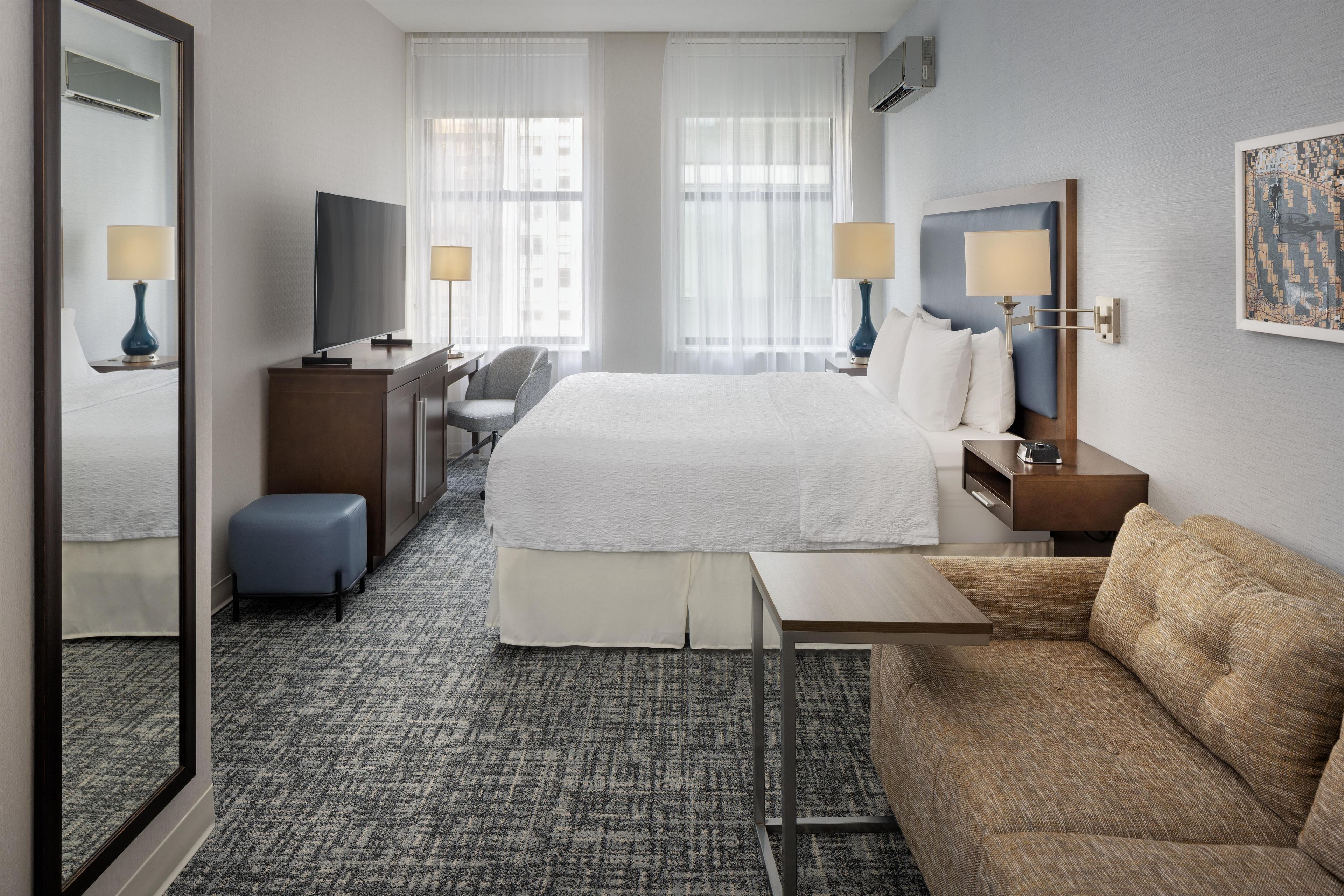 Quarto de hotel com cama, escrivaninha, poltrona, sofá, televisão e espelho.