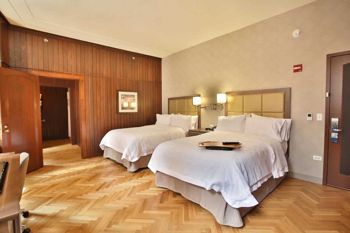 Une chambre d’hôtel avec deux lits, des lambris en bois aux murs, un parquet en bois disposé en chevrons, un bureau et une chaise.