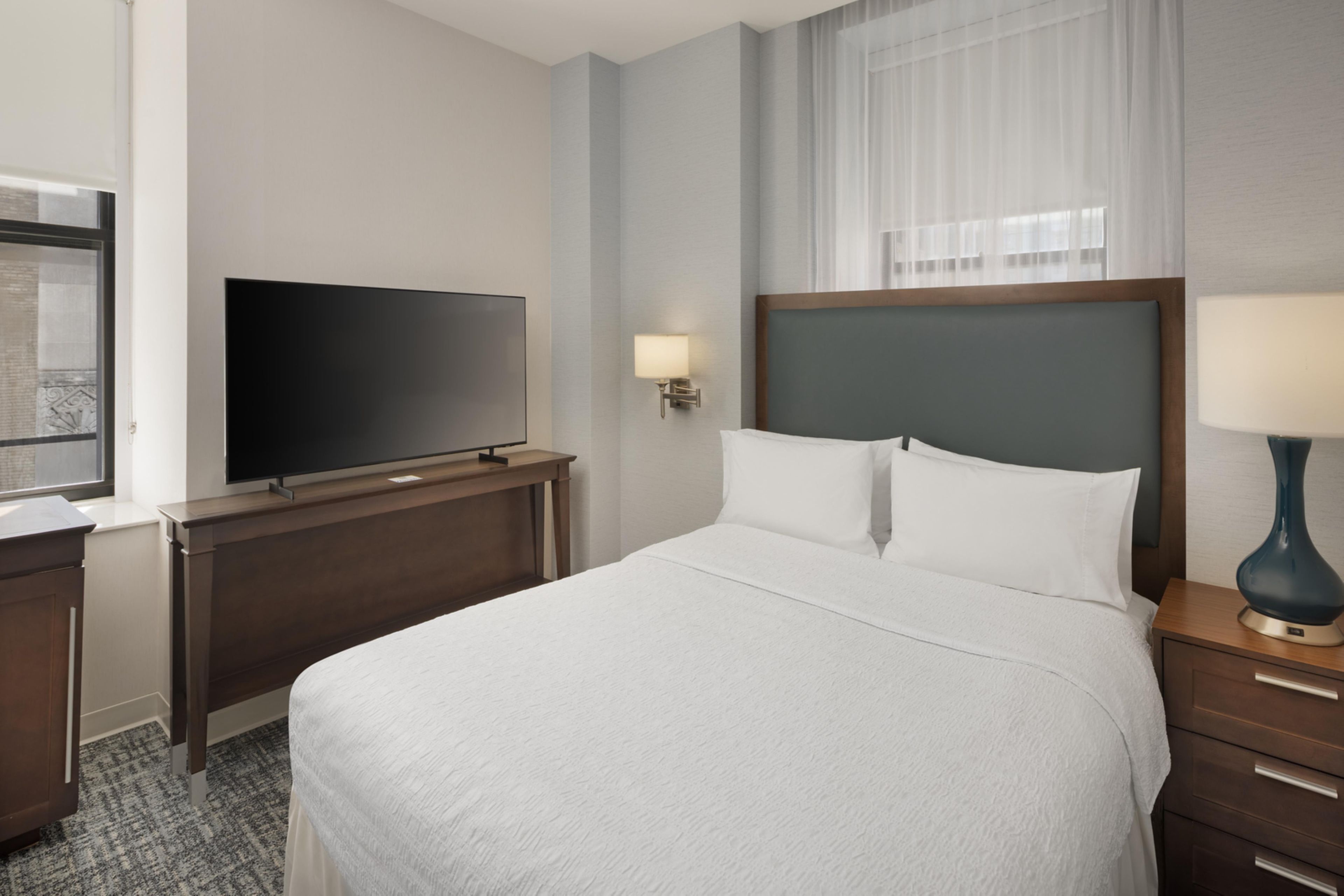 Une chambre d’hôtel avec un lit, un téléviseur posé sur un meuble, une table de chevet avec une lampe et une fenêtre avec des rideaux légers.