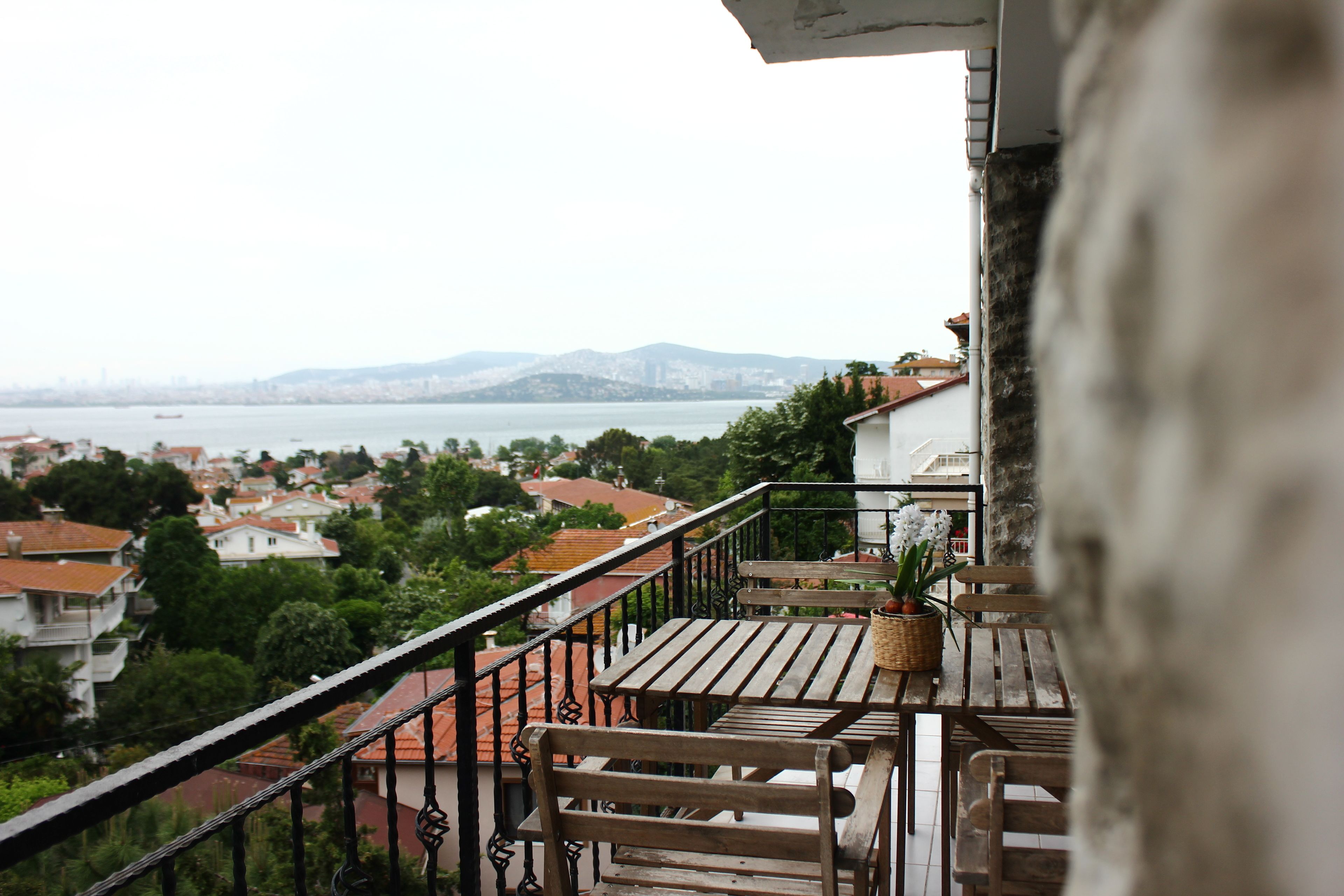 A balcony with a wooden table and chairs, overlooking a cityscape with buildings and a body of water.